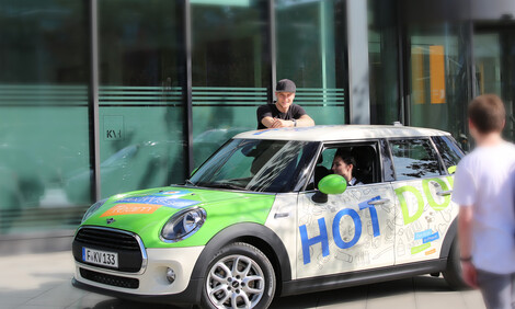 Ein Auto mit der Aufschrift Hot Doc steht vor dem KVH-Gebäude, am Steuer sitzt eine junge Frau. Ein junger Mann lehnt sich aufs Dach, ein anderer läuft auf das Auto zu mit dem Rücken zur Kamera.