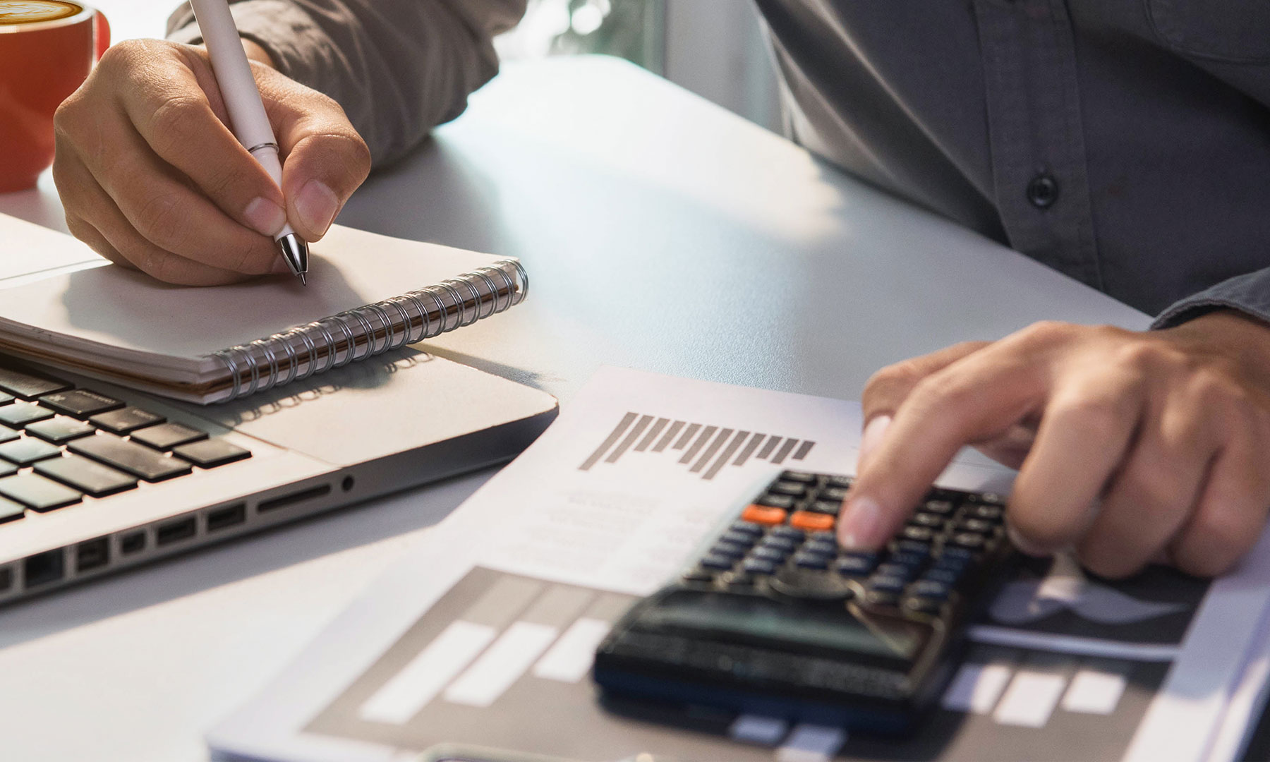 Ein Mann sitzt vor seinem Laptopt, tippt mit einer Hand etwas in den Taschenrechner und mit der anderen schreibt er etwas auf ein Notizblock.