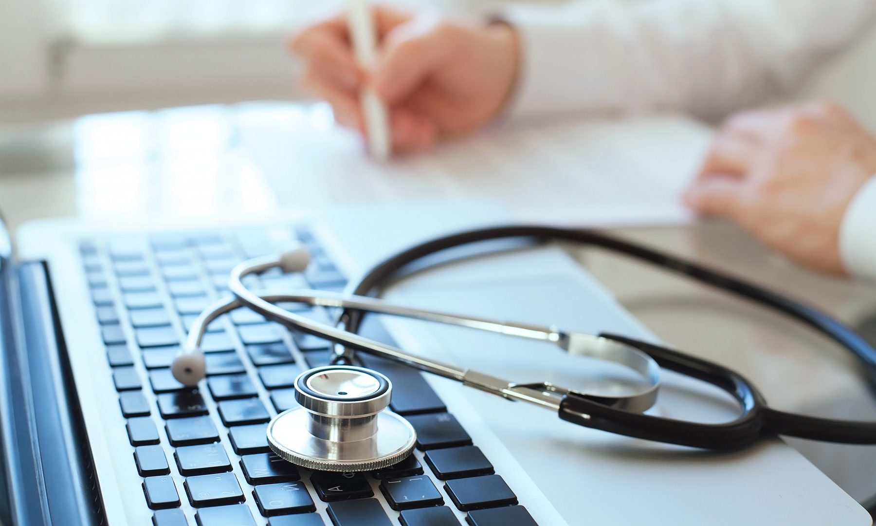 Ein Stethoskop liegt auf einer Laptop-Tastatur. Im Hintergrund sitzt ein Arzt am Schreibtisch und notiert etwas auf einem Zettel. 